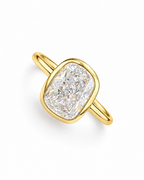 The Tilde Bezel Ring in polished yellow-gold tone with one clear faceted rounded-rectangle stone in a diagonal pearl-white studio flat lay — reference-based AI visualization