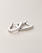 The Aria CZ Hoops — overlapping polished silver-colored U-shaped hoops with clear round stone rows on pale greige, reference-based AI visualization