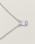The Mara Geometric CZ Pendant — jewelry-only lateral layout with the horizontal clear rectangular stone, four corner prongs, side rings, and alternating-link chain on pale warm greige; reference-based AI visualization