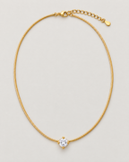 Gold-color necklace arranged as a complete vertical closed loop with a clear round four-prong stone below and clasp with extension above on pale warm greige; reference-based AI visualization.