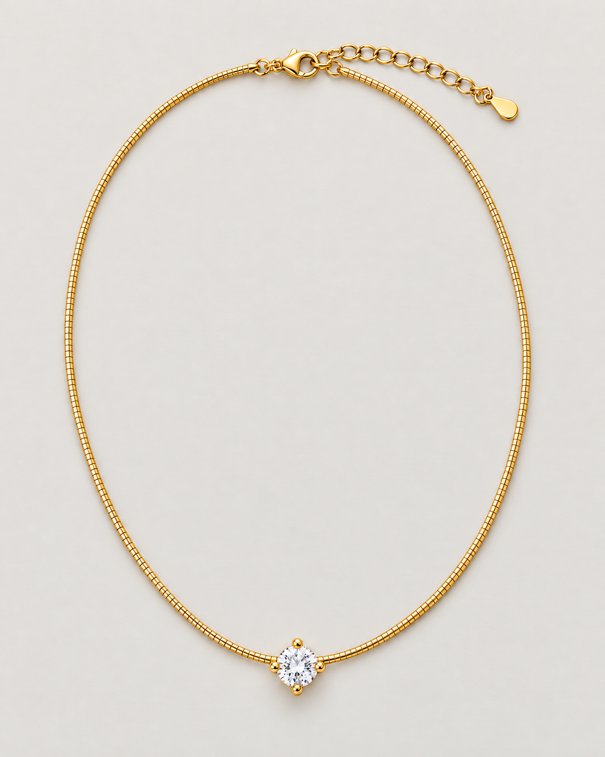 Gold-color necklace arranged as a complete vertical closed loop with a clear round four-prong stone below and clasp with extension above on pale warm greige; reference-based AI visualization.