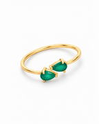 The Tove Green Twin-Pear Ring in polished yellow-gold coloured metal with two green pear-cut stones in a diagonal flat lay on a pearl-white studio background, reference-based AI visualization.