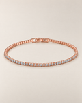The Mila Tennis Bracelet in Rose Gold Tone, shown as a full oval with clear stones and a closed clasp on a warm pale-greige studio background, reference-based AI visualization.