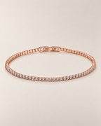 The Mila Tennis Bracelet in Rose Gold Tone, shown as a full oval with clear stones and a closed clasp on a warm pale-greige studio background, reference-based AI visualization.