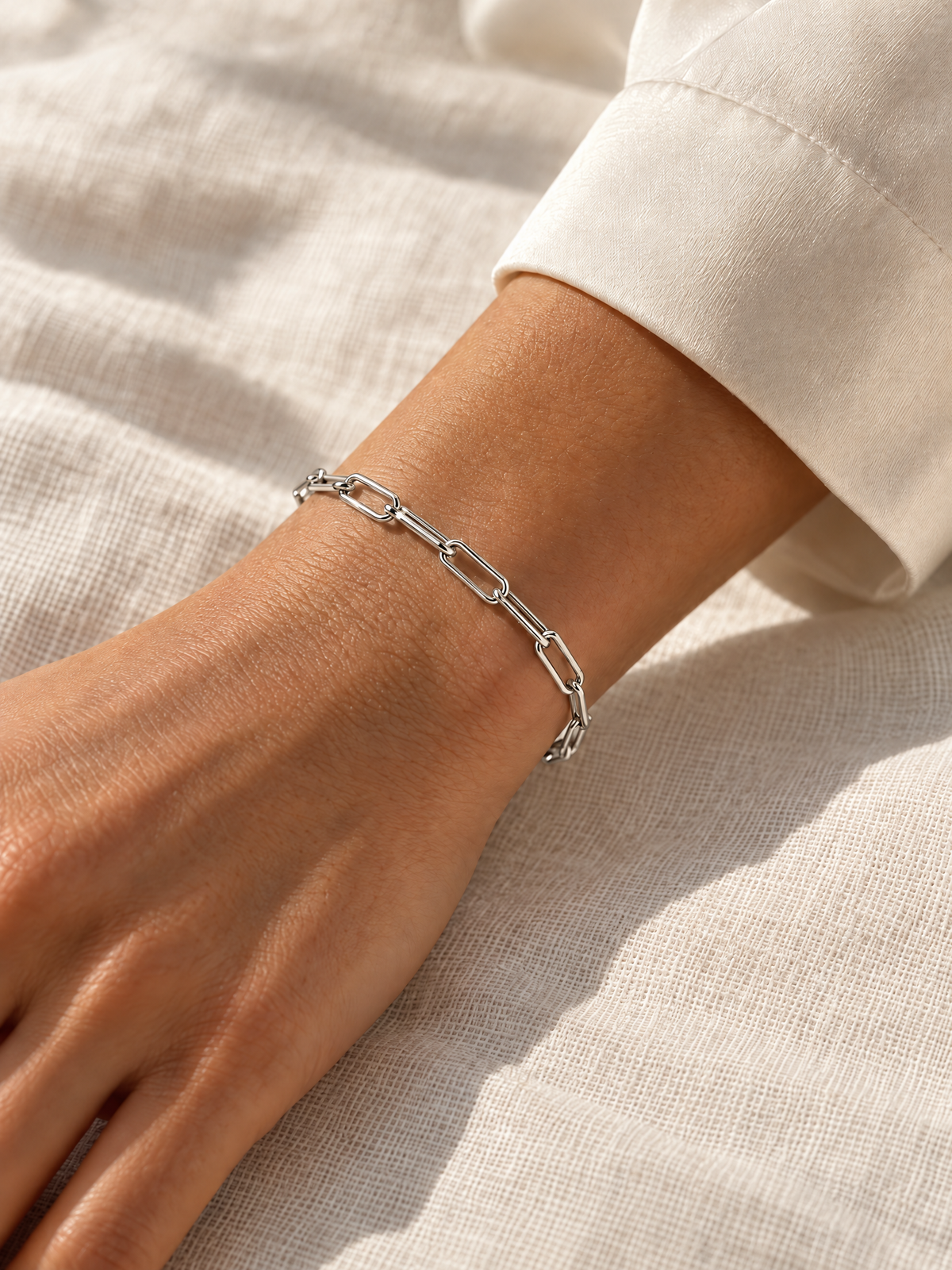 Silver-tone Aelin Paperclip Bracelet on a wrist over ivory linen in matching warm side light — reference-based AI lifestyle image.
