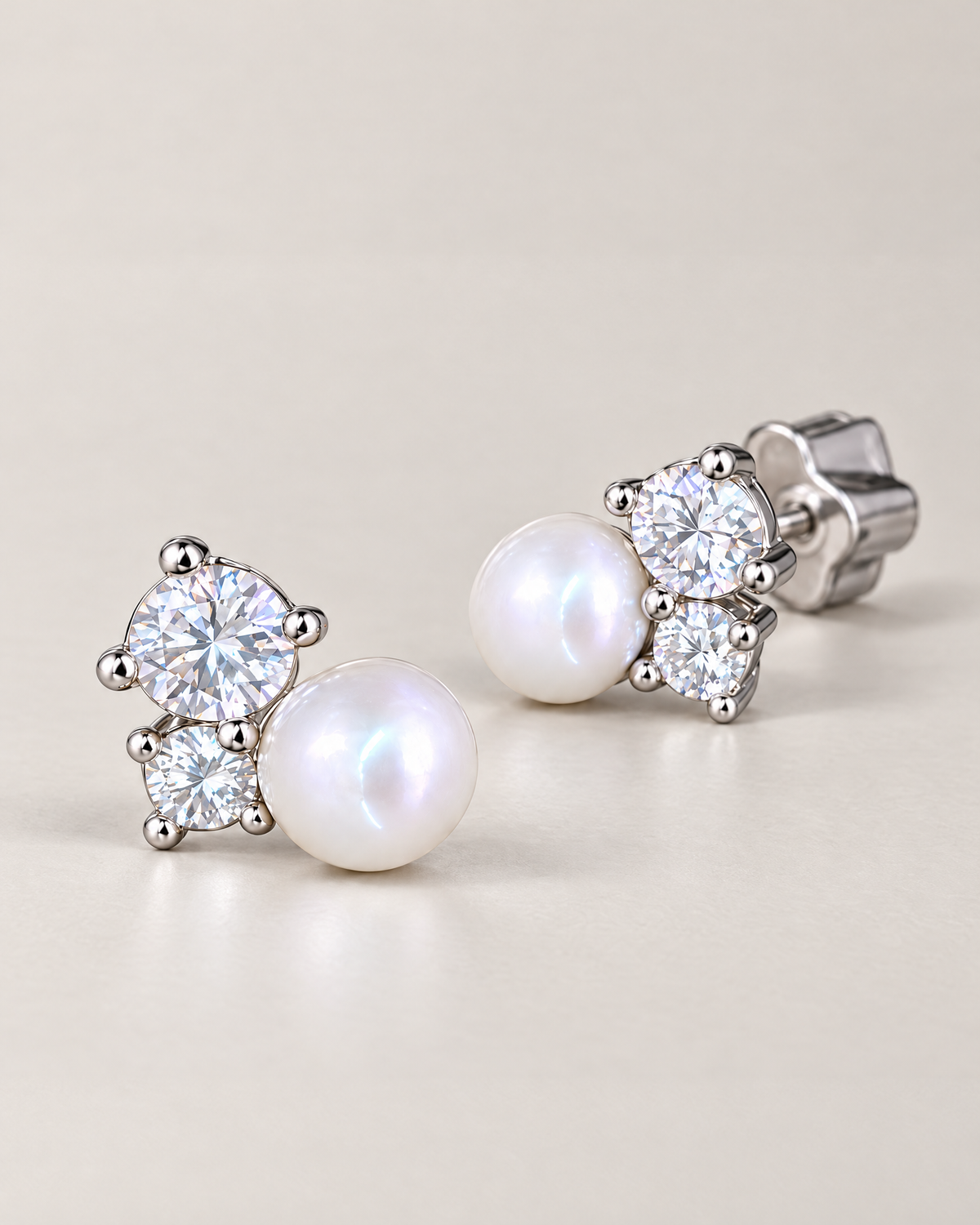 The Elva Pearl & Moissanite Stud Earrings, low-angle pair of silvery studs with white pearls, clear round stones and one scalloped butterfly back on pale greige, reference-based AI visualization.