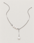The Freya Pearl Drop Necklace — cool silver-color cable chain in a relaxed open-U lay with three white pearl-like accent beads and a single pearl-like drop on pale greige, reference-based AI visualization