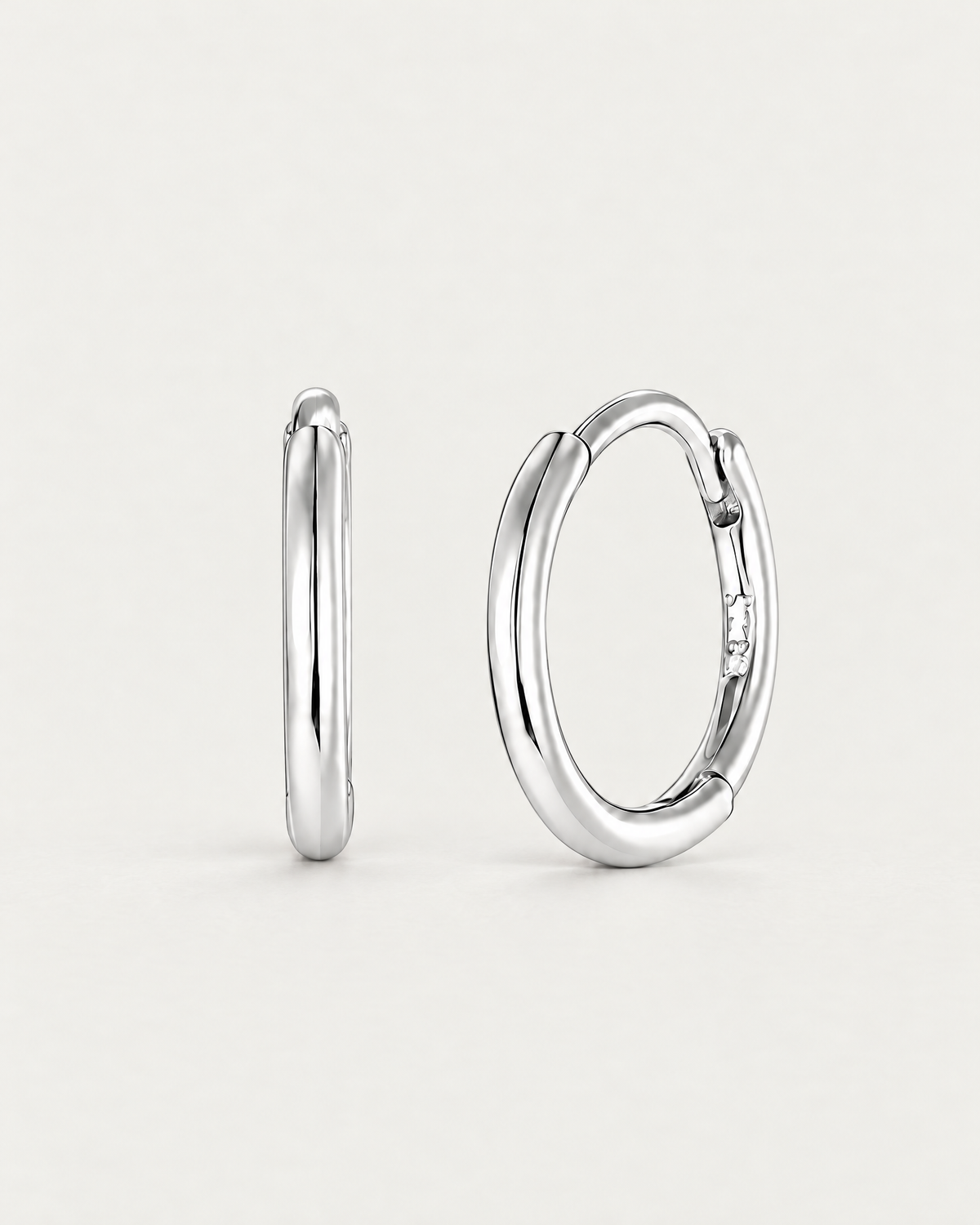 The Kiri Huggie Hoops — upright silver pair, front/edge-on and three-quarter views on pale greige; reference-based AI visualization.