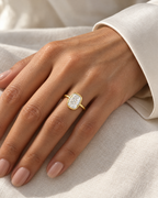 The Tilde Bezel Ring in polished yellow-gold tone with one clear faceted rounded-rectangle stone, correctly worn on a medium-warm adult hand against ivory fabric — reference-based AI visualization
