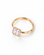 The gold-finish Eira Square Solitaire Ring shown complete at an overhead resting angle on pearl white with its single clear square stone and smooth bezel; reference-based AI visualization.