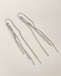 The Neri Threader Earrings, paired bright silver-tone threaders in a diagonal overhead arrangement on pale greige, reference-based AI visualization