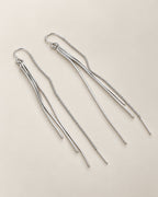 The Neri Threader Earrings, paired bright silver-tone threaders in a diagonal overhead arrangement on pale greige, reference-based AI visualization