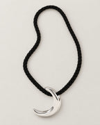 Black braided cord in a tall asymmetric loop with a polished crescent pendant at its base on pale greige, reference-based AI visualization