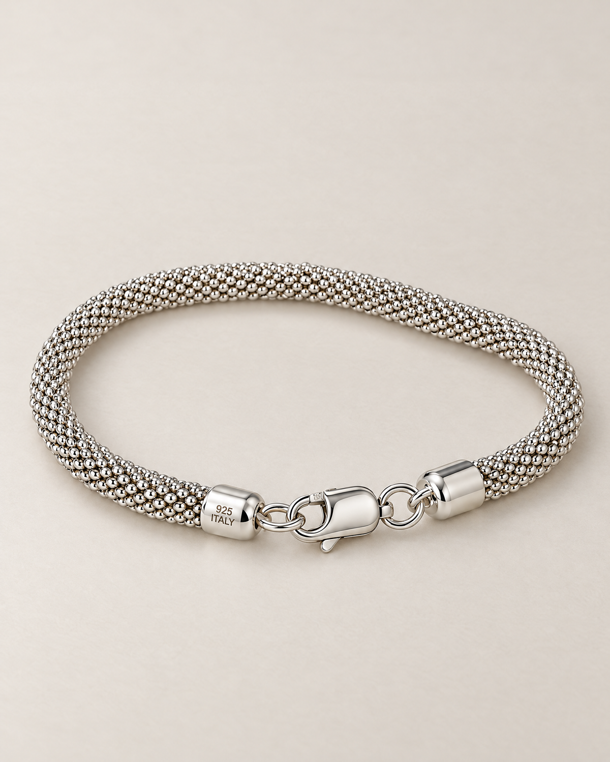 The Liva Mesh Chain Bracelet in a complete low resting loop of silver-tone woven mesh with lobster clasp and visible 925 ITALY end-cap marking on warm pale greige; reference-based AI visualization.