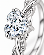 The Alva Heart-Stone Ring, observed polished silver-tone, macro detail of the clear heart-shaped center stone, four rounded prongs, and twisted beaded shoulder on a pearl-white studio background — reference-based AI visualization.