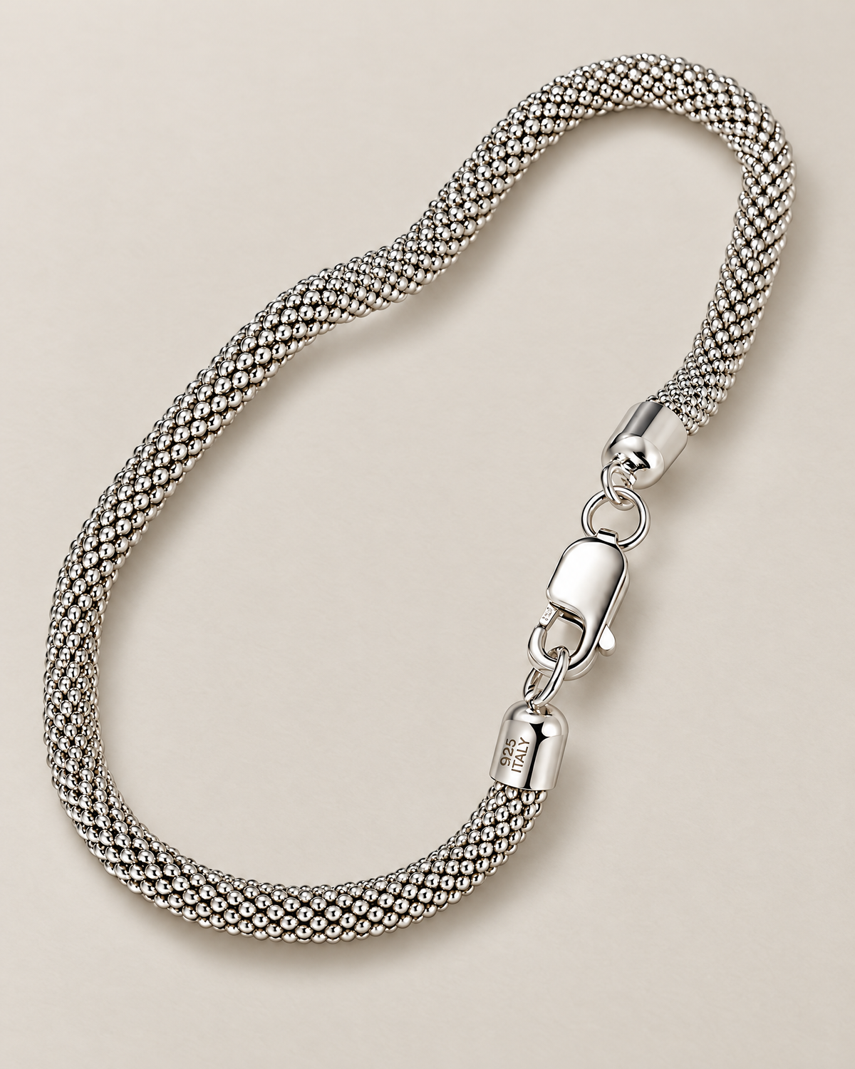 The Liva Mesh Chain Bracelet in silver-tone woven mesh, shown as a full three-quarter loop with lobster clasp and visible 925 ITALY end-cap marking on warm pale greige; reference-based AI visualization.