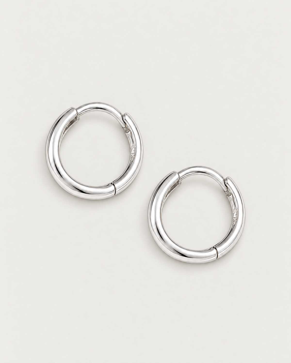 The Kiri Huggie Hoops — silver pair — separated overhead lay on pale greige, product-only; reference-based AI visualization.