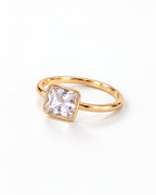 The gold-finish Eira Square Solitaire Ring shown complete at a low flat-lay angle on pearl white, with the single square stone toward the left; reference-based AI visualization.