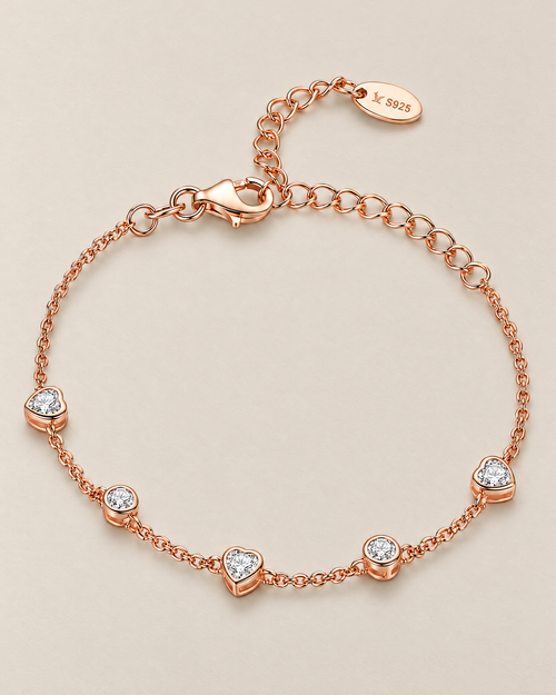 The Dahlia S925 Heart Bracelet in warm rose-gold-toned metal with five alternating clear heart and round stone stations in a full front oval on pale greige, reference-based AI visualization.