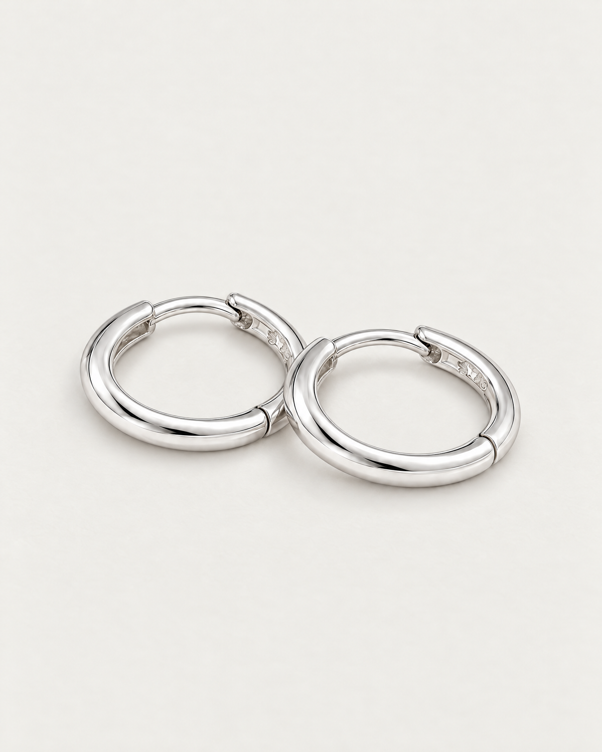The Kiri Huggie Hoops — silver pair — low-angle overlapping lay on pale greige, product-only; reference-based AI visualization.