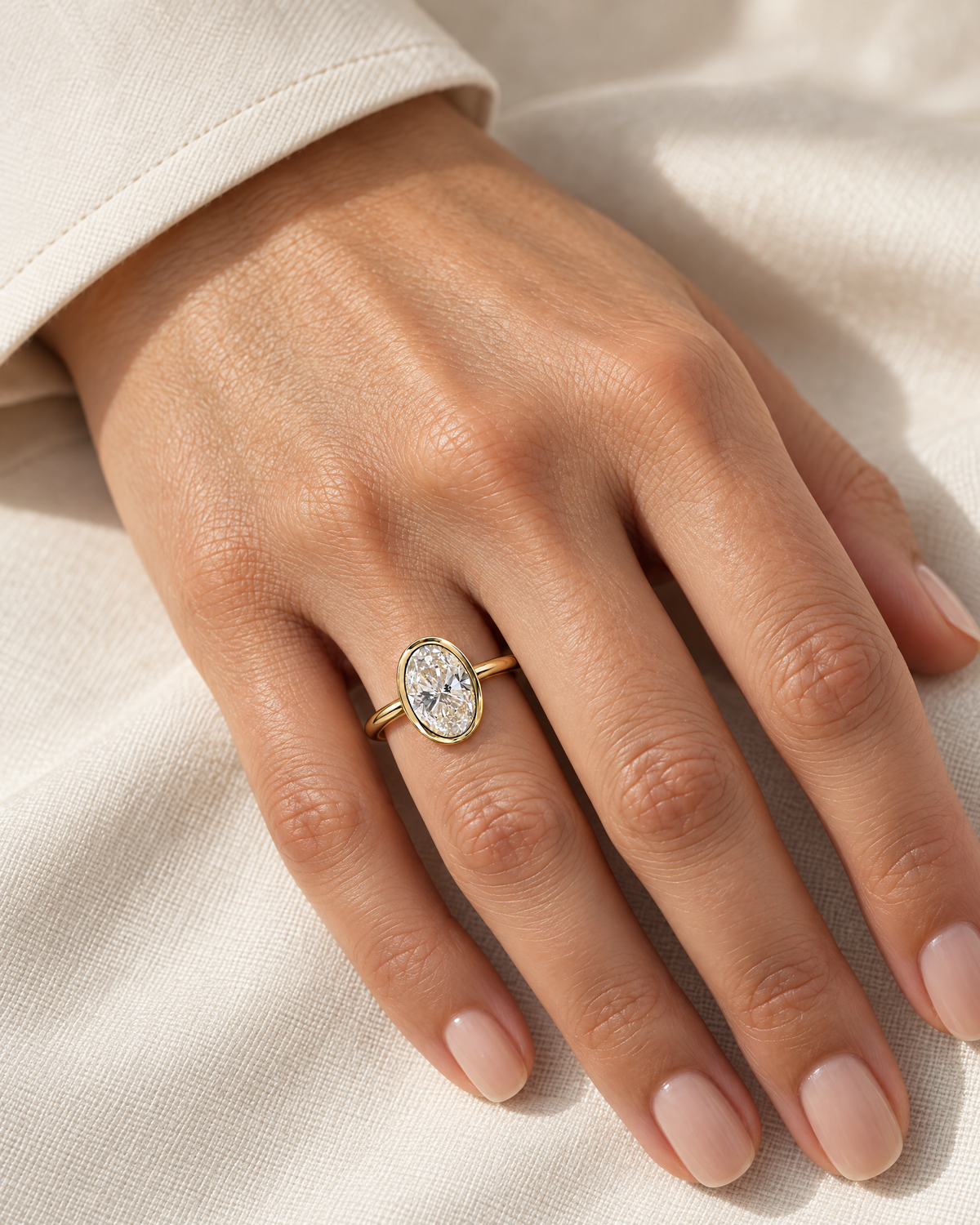 The Esme Bezel-Set Oval Ring in Gold Tone, elegant on-hand view on a light-warm adult hand with ivory linen, reference-based AI visualization.