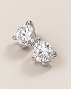 The Elio Moissanite Stud Earrings — overlapping clear round faceted studs in silver-colored prong settings on pale greige, reference-based AI visualization