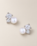The Elva Pearl & Moissanite Stud Earrings, separated overhead pair of silvery studs with white pearls and clear round stones on pale greige, reference-based AI visualization.