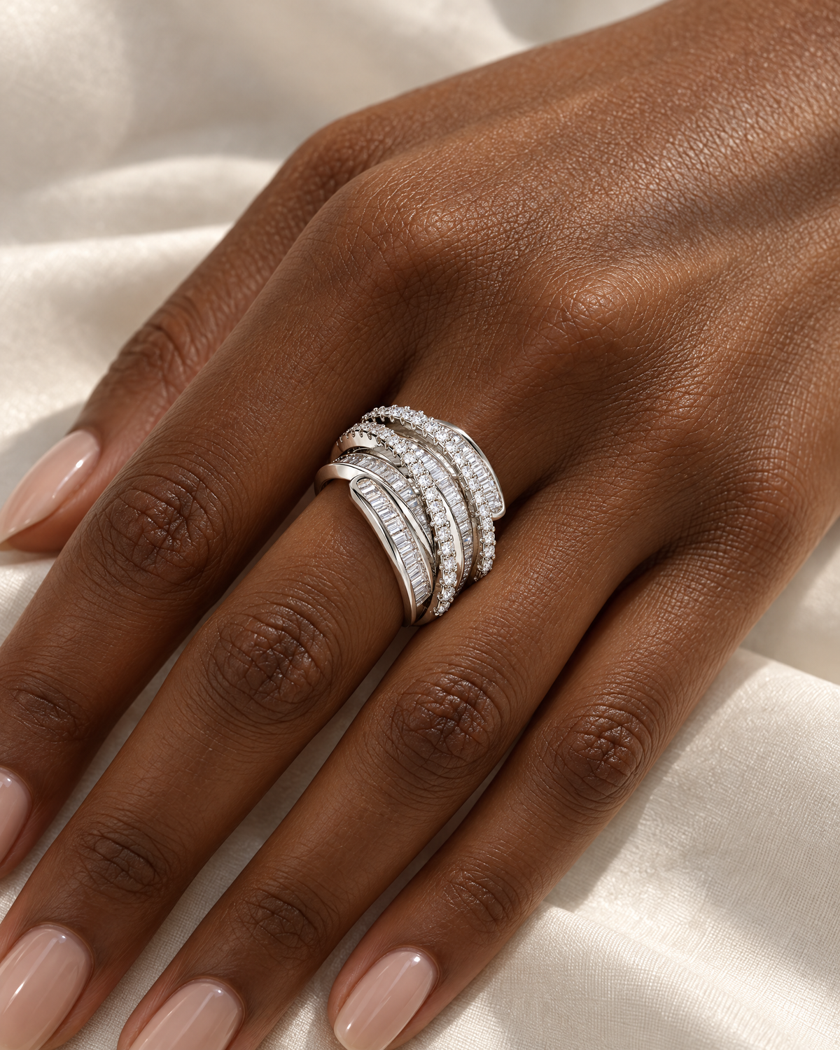The Iona Interwoven Ring — White/silver-tone interwoven ring worn on a deep brown-skinned adult hand over ivory satin; reference-based AI visualization.