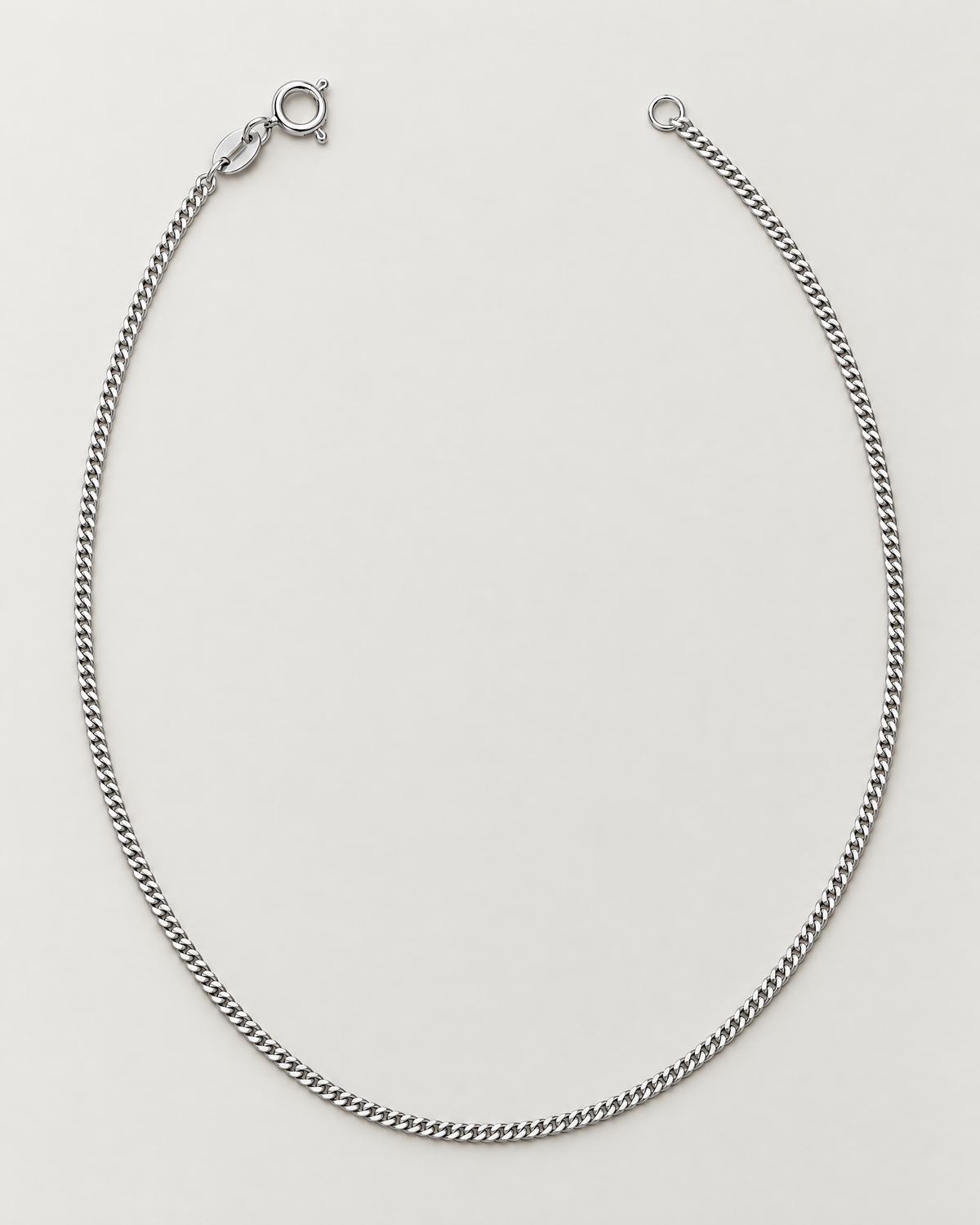 The Erin Fine Curb Chain Necklace — Sterling Silver complete elongated open-U curb-chain hero with spring-ring clasp and oval tag on pale greige, reference-based AI visualization.