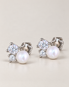 The Elva Pearl & Moissanite Stud Earrings, matching silvery studs with luminous white pearls and two clear round stones per earring in a front three-quarter pair composition on pale greige, reference-based AI visualization.