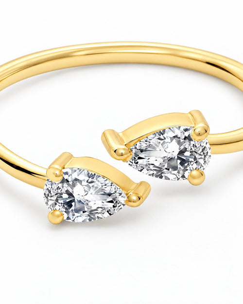The Agnes Twin-Pear Ring in a polished yellow-gold finish with two clear pear-shaped stones, front setting macro on a pearl-white studio background — reference-based AI visualization