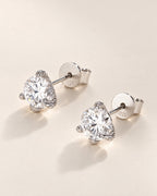 The Elio Moissanite Stud Earrings — clear round faceted studs in silver-colored prong settings arranged diagonally with posts and circular S925-marked backs on pale greige, reference-based AI visualization