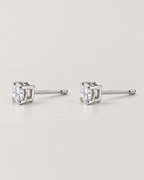 The Neva CZ Stud Earrings — silver-tone side-profile pair showing open settings, clear round stones, and straight posts in a pale-greige studio; reference-based AI visualization.