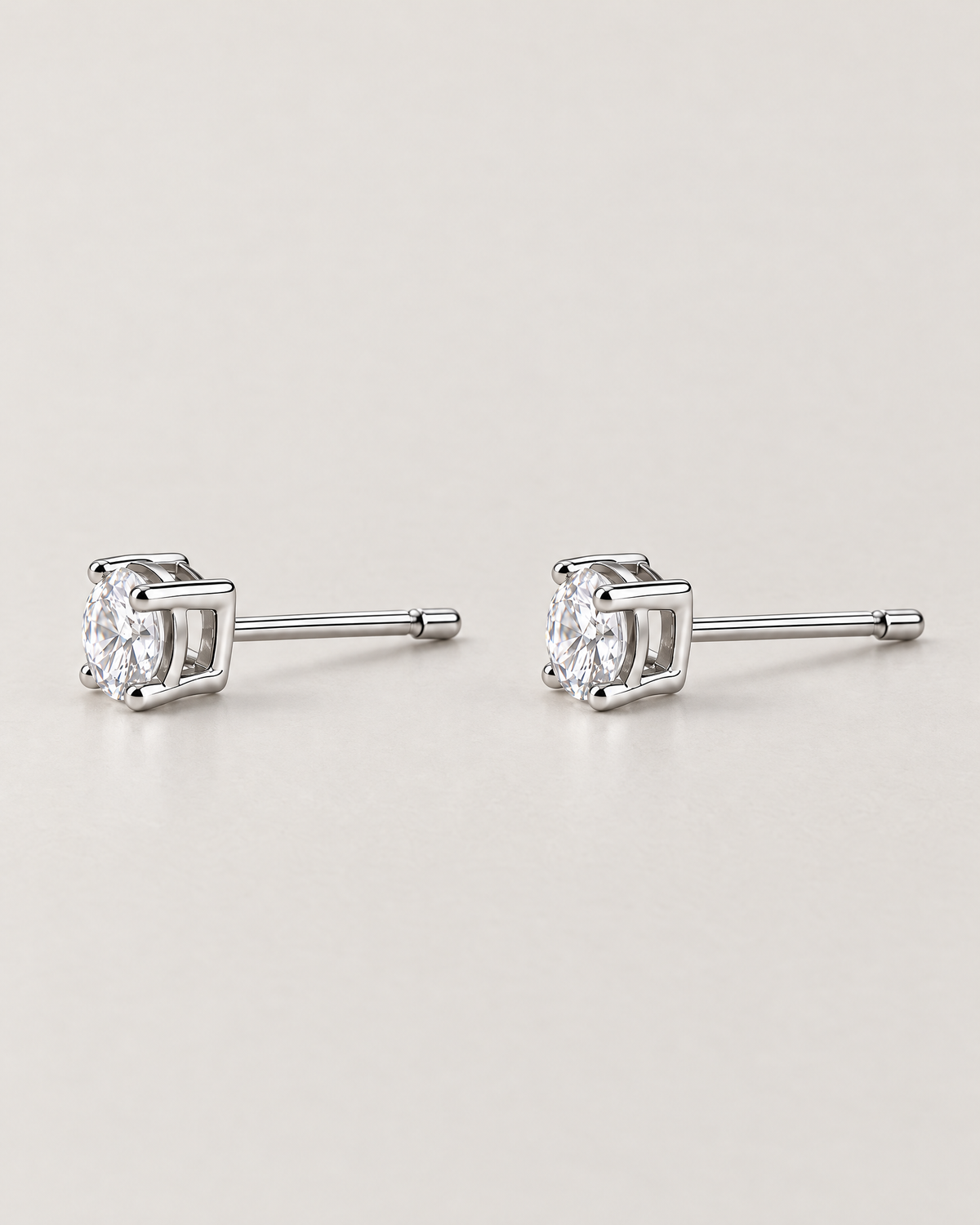 The Neva CZ Stud Earrings — silver-tone side-profile pair showing open settings, clear round stones, and straight posts in a pale-greige studio; reference-based AI visualization.