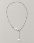 The Freya Pearl Drop Necklace — cool silver-color cable chain with three white pearl-like accent beads and a single pearl-like Y-drop on pale greige, reference-based AI visualization