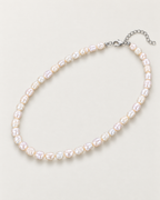 Irregular off-white pearl strand in a complete diagonal oval with clasp and extension at upper right on a pale greige studio surface; reference-based AI visualization