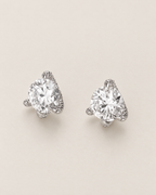 The Elio Moissanite Stud Earrings — two separated clear round faceted studs in silver-colored prong settings viewed overhead on pale greige, reference-based AI visualization