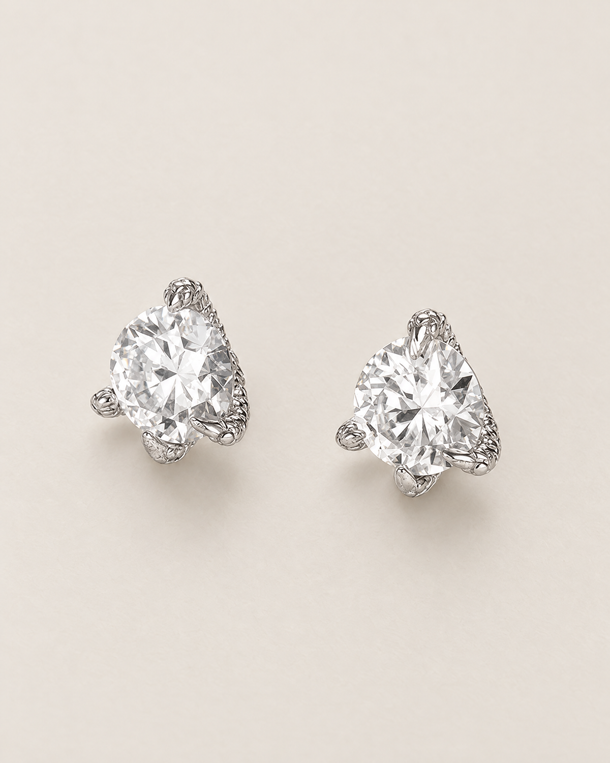 The Elio Moissanite Stud Earrings — two separated clear round faceted studs in silver-colored prong settings viewed overhead on pale greige, reference-based AI visualization
