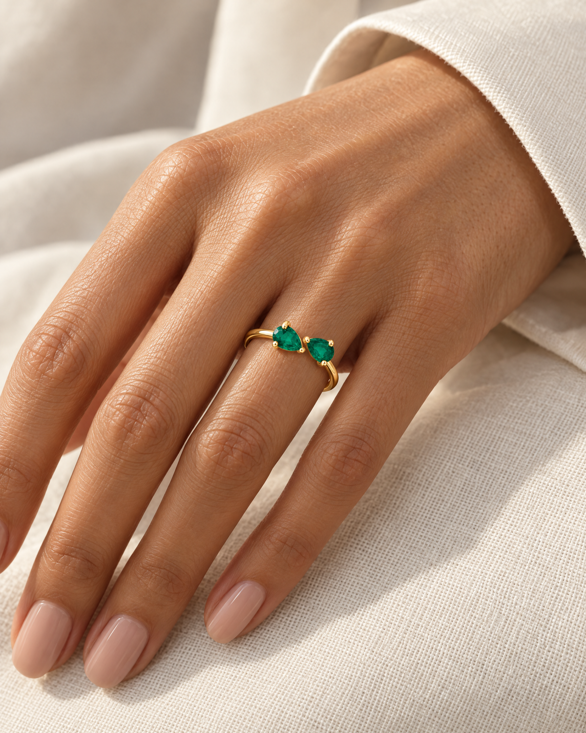 The Tove Green Twin-Pear Ring in polished yellow-gold coloured metal with two green pear-cut stones worn on a medium-warm adult hand with an ivory linen cuff, reference-based AI visualization.