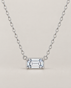 The Mara Geometric CZ Pendant — jewelry-only straight-on hero with a horizontal clear rectangular stone, four corner prongs, side connector rings, and an alternating-link chain on pale warm greige; reference-based AI visualization