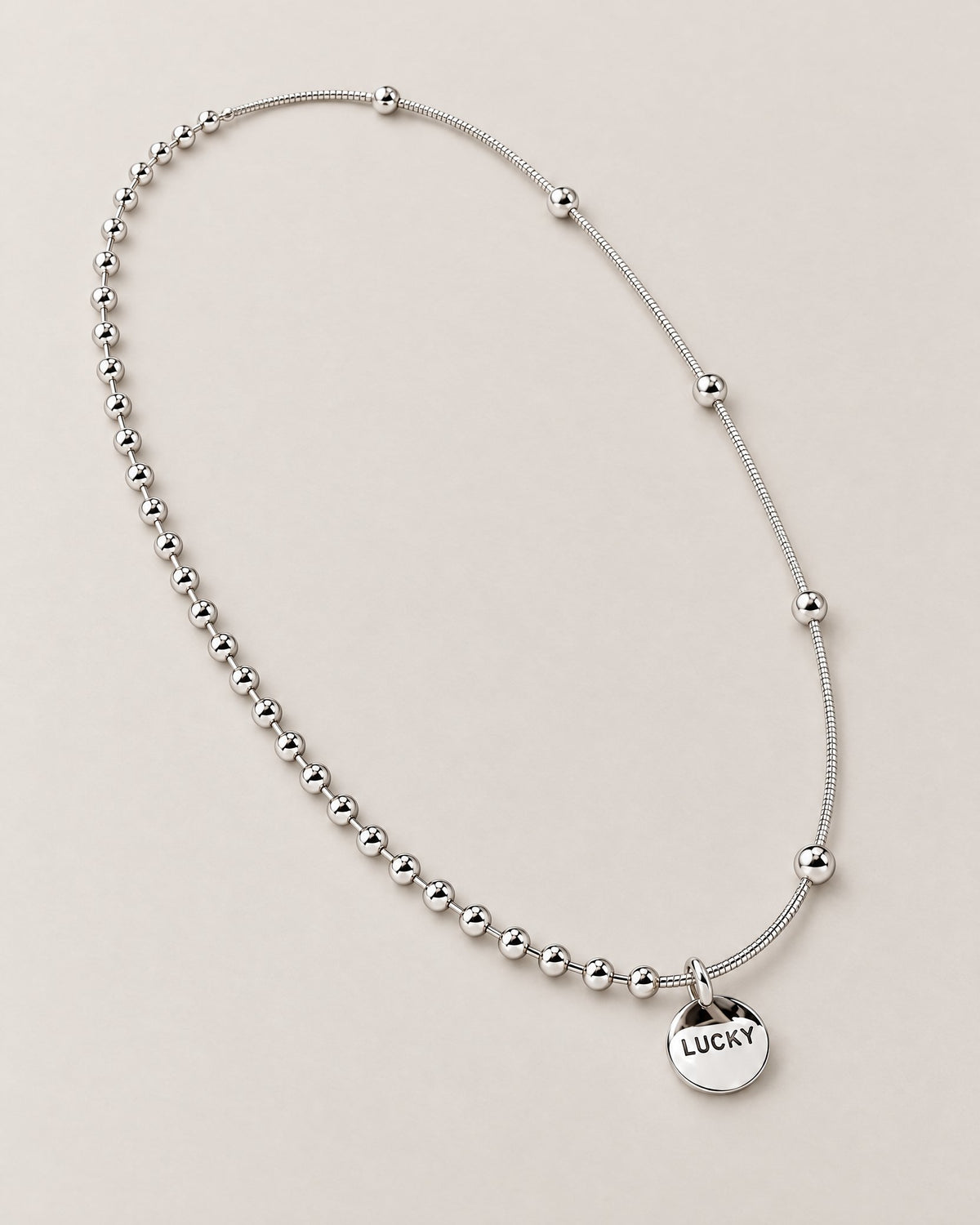 Diagonal overhead oval lay of the silver-colored asymmetric necklace with a front LUCKY medallion and contrasting beaded and smooth station-bead chain sections, reference-based AI visualization