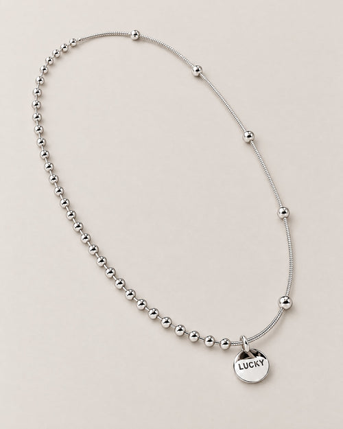Diagonal overhead oval lay of the silver-colored asymmetric necklace with a front LUCKY medallion and contrasting beaded and smooth station-bead chain sections, reference-based AI visualization