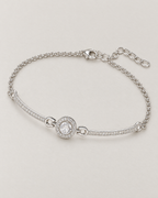 The Sora Halo Bracelet polished cool silver-tone complete diagonal overhead lay with clear-stone halo, curved stone-set bars and chain extension on pale greige reference-based AI visualization