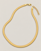 Gold-tone flat herringbone necklace in an asymmetrical open loop with the clasp and oval-link extender separated at the top on a pale greige studio ground; reference-based AI visualization
