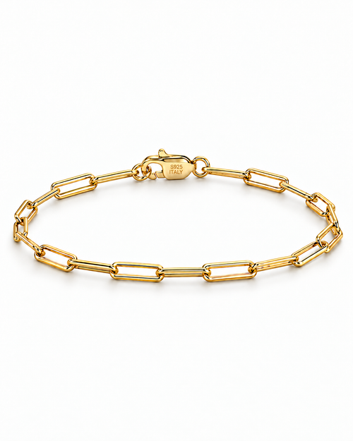The Aelin Paperclip Bracelet in polished yellow-gold tone, shown as a full closed oval with the marked clasp at the top on a plain pearl-white studio surface; reference-based AI visualization