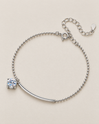 The Eden Moissanite Bracelet in polished silver tone, shown in a relaxed overhead curve with the curved bar, one round clear stone, ball-bead chain, clasp, and extension on a pale-greige background, reference-based AI visualization.