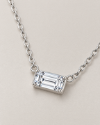 The Mara Geometric CZ Pendant — jewelry-only three-quarter view of the horizontal clear rectangular stone, four corner prongs, side rings, and alternating-link chain on pale warm greige; reference-based AI visualization