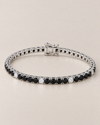 The Signe Black & White CZ Tennis Bracelet in polished silver-tone metal, shown in a low resting oval with repeating black and clear/white round stones and a box clasp on pale greige, reference-based AI visualization.