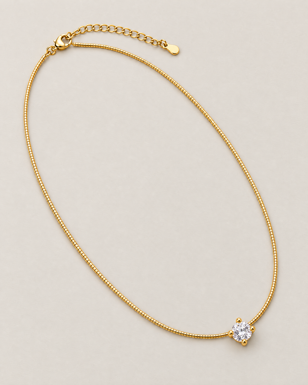 Gold-color necklace in a complete diagonal oval lay with its clear round four-prong stone, lobster clasp, extension links, and smooth end tag on pale warm greige; reference-based AI visualization.