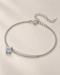 The Eden Moissanite Bracelet in polished silver tone, arranged as a full loop with a curved bar, one round clear stone, ball-bead chain, and extension clasp on a pale-greige background, reference-based AI visualization.